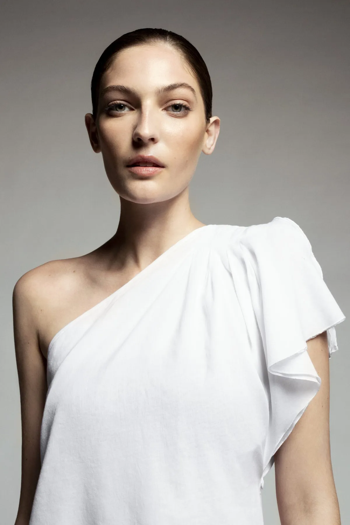 Model in a white one-shoulder top with a ruffled sleeve, hair pulled back, looking to camera