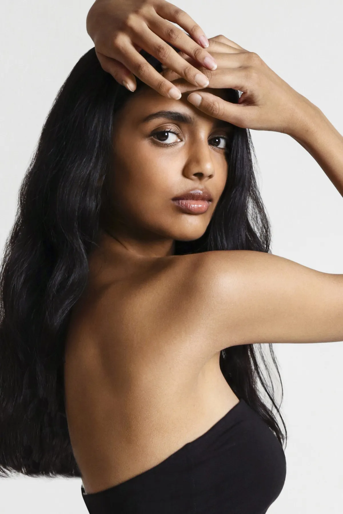 Model in a black strapless top, both hands resting on top of her head, long hair over one shoulder