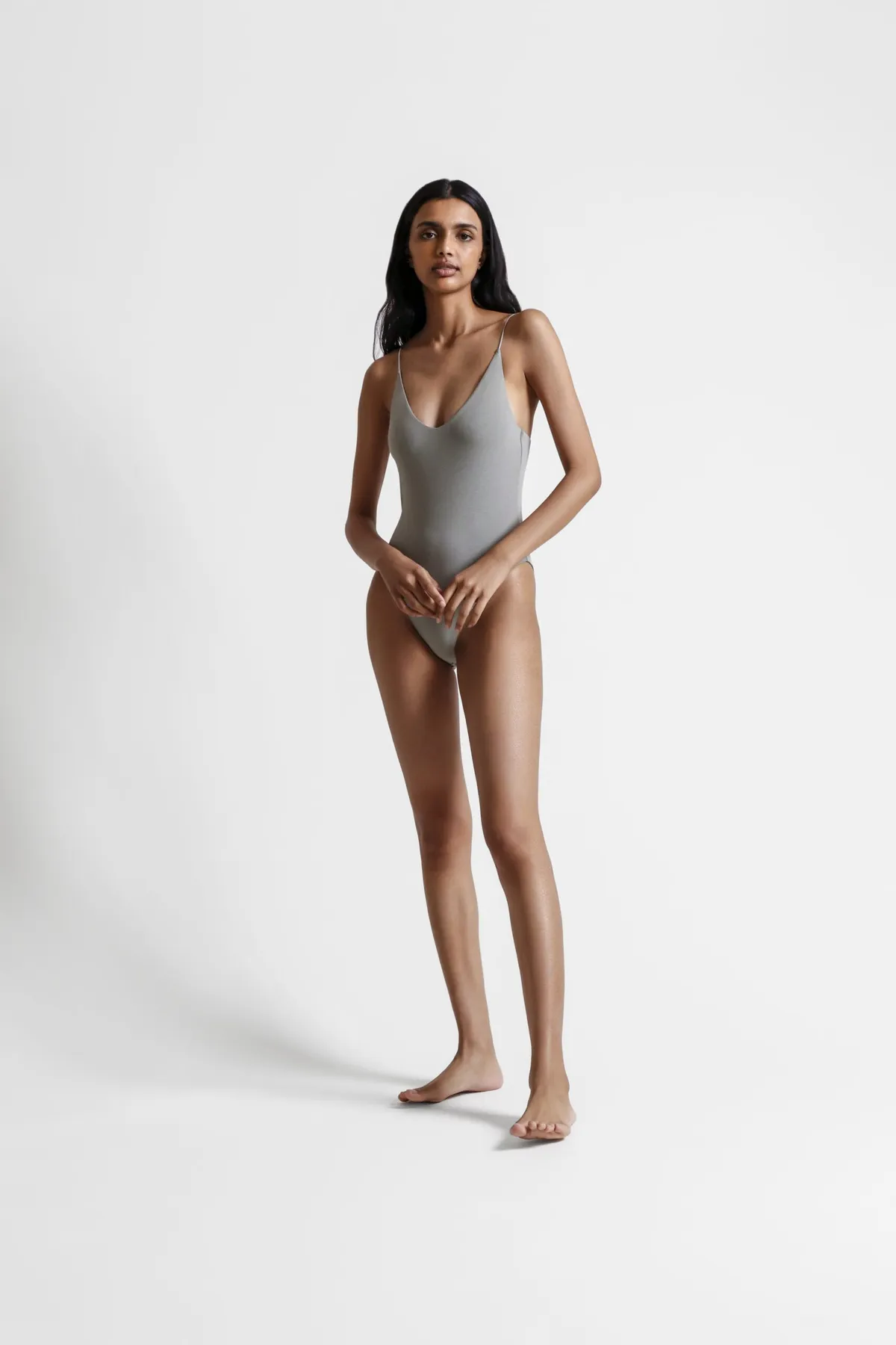 Full-length: model in a sage bodysuit, hands clasped low, on a white ground