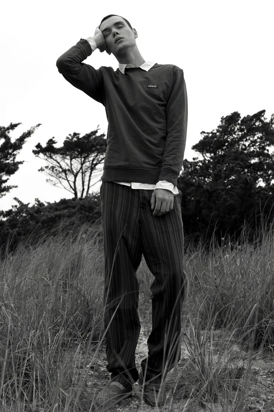 Black and white: man in a dark polo and striped trousers standing in dune grass, hand to his head