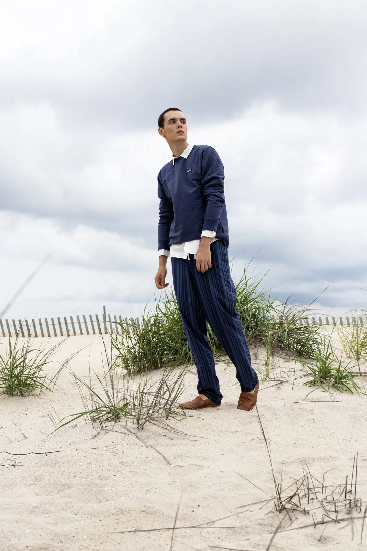 Man in a navy top and striped trousers standing on a sand dune under a cloudy sky