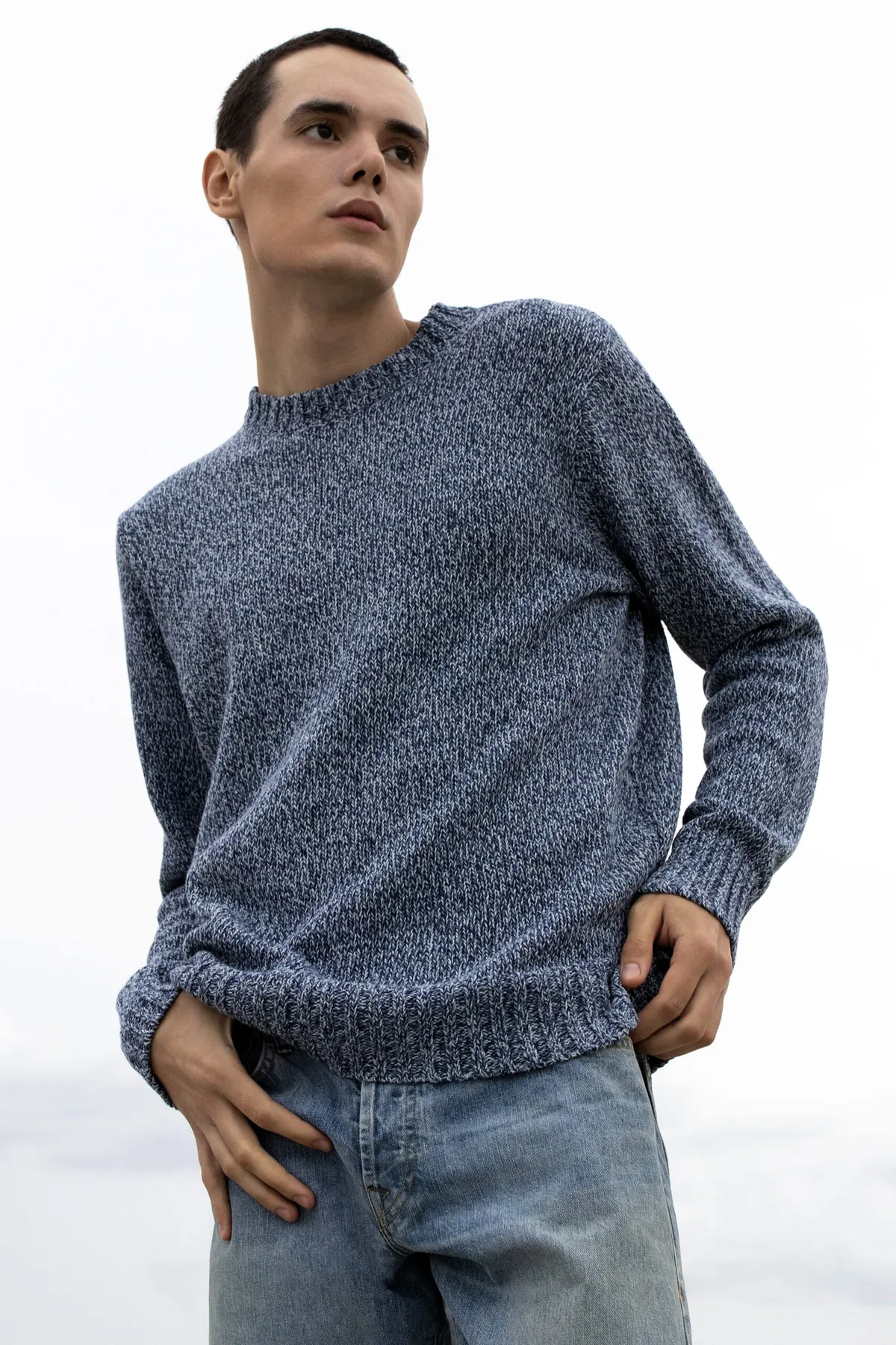 Man in a marled blue sweater and jeans, hand in pocket, looking up against a pale sky