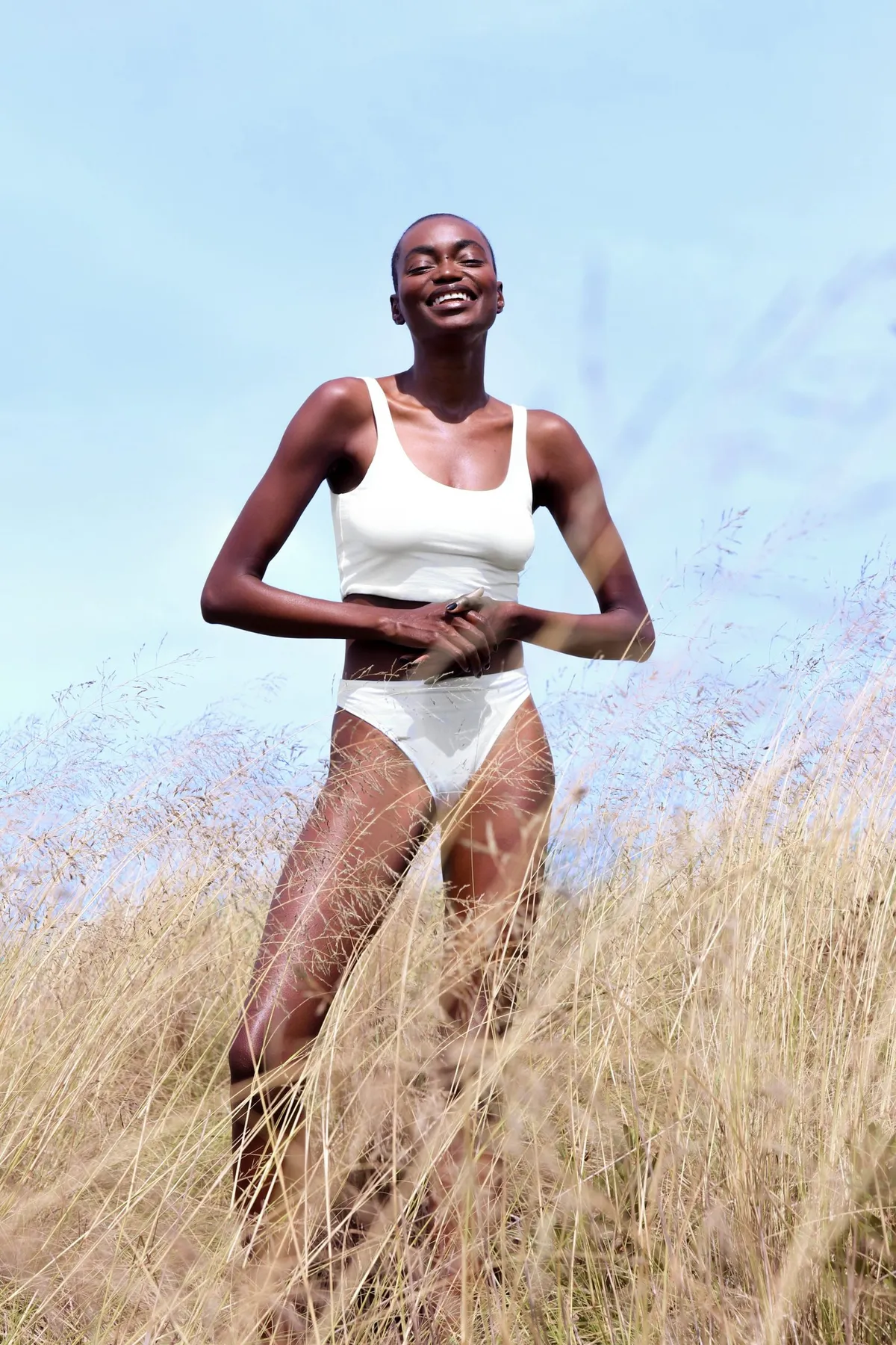 Model in a white two-piece standing in tall golden grass under a blue sky, eyes closed to the sun