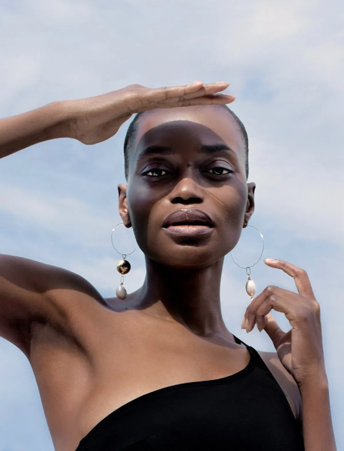 Model in a black one-shoulder swimsuit shielding her eyes, hazy blue sky behind