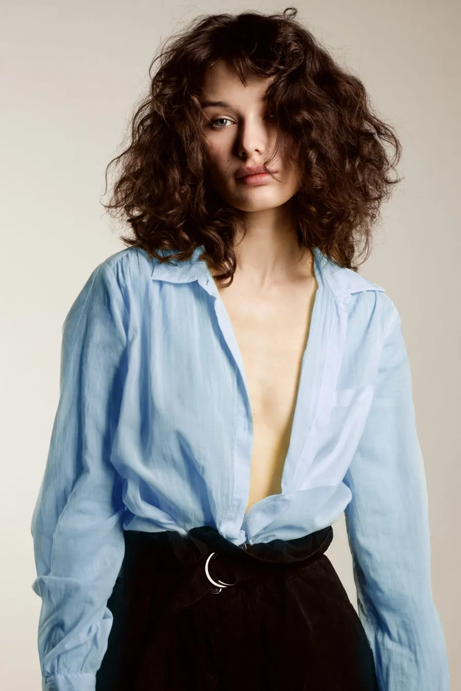 Model in an open pale blue shirt and black belted skirt, curly hair over one eye