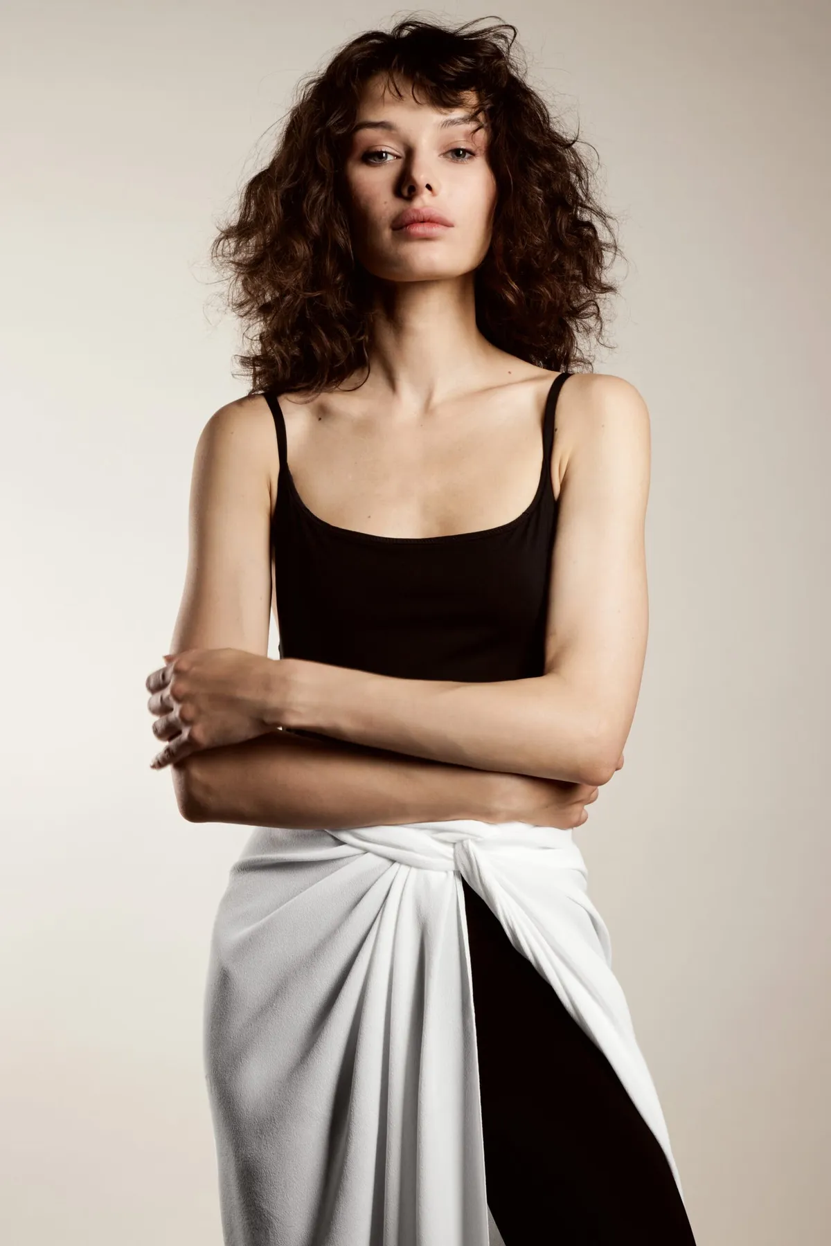 Model in a black camisole and white wrap skirt, arms folded, looking to camera