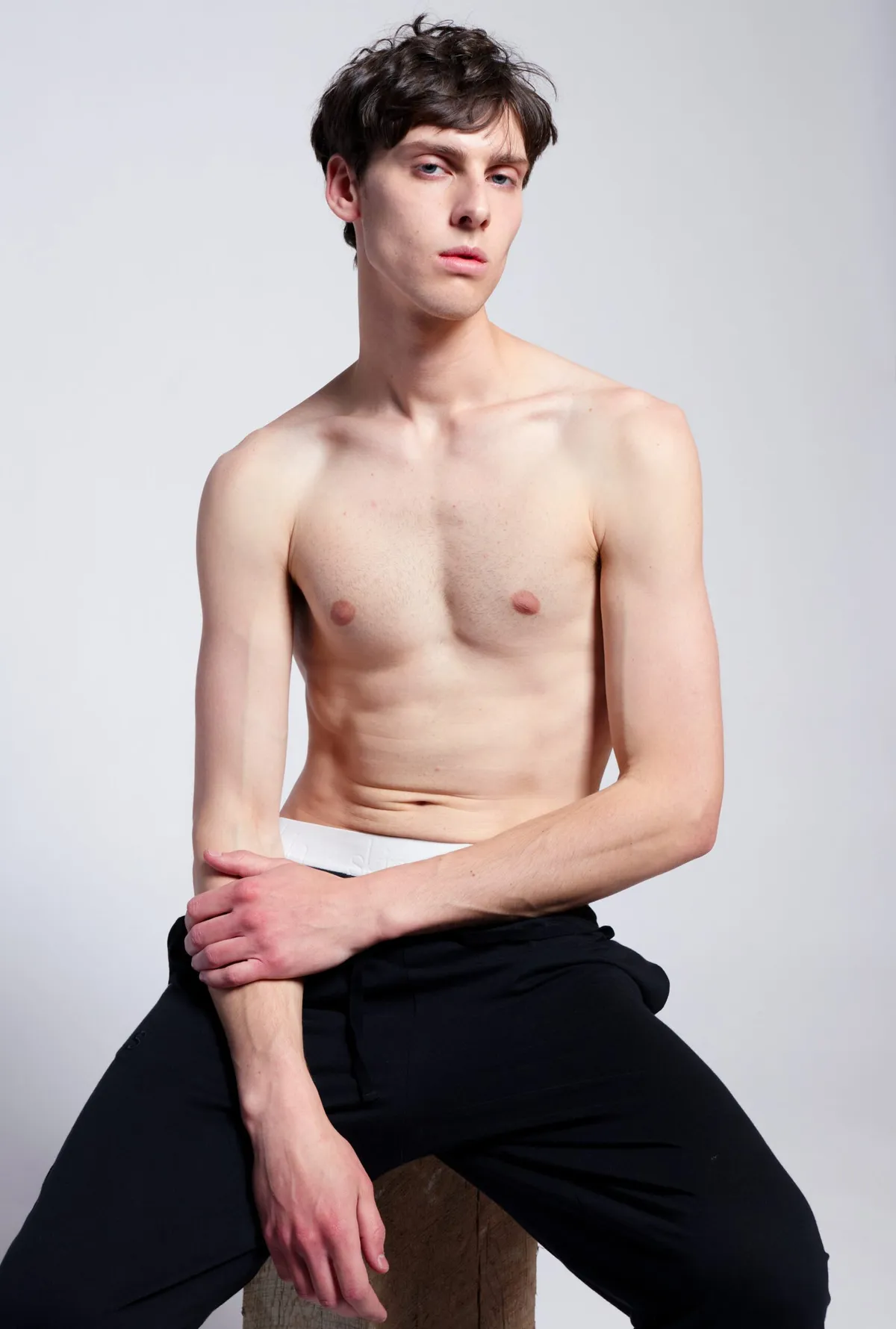 Shirtless man in black trousers seated on a stool, arms crossed over his knee